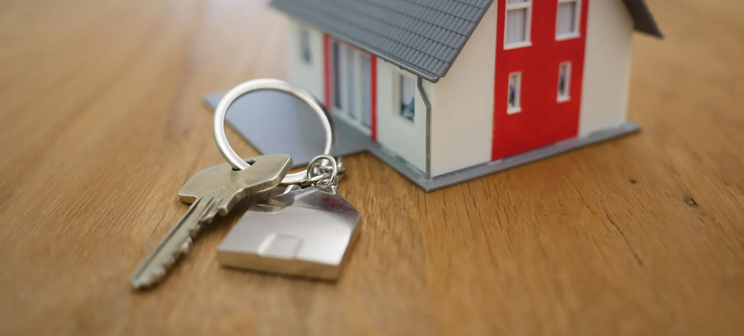 Ein Haustürschlüssel mit Hausanhänger liegt vor einem Modellhaus auf einem Holztisch
